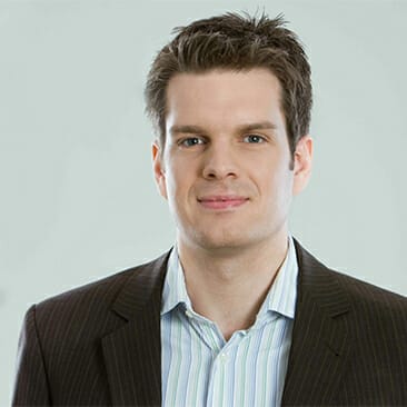 A man with short, dark hair is wearing a striped shirt and a dark pinstriped suit jacket. He is facing the camera with a neutral expression against a plain light background, conveying the poised professionalism typical in digital commerce.