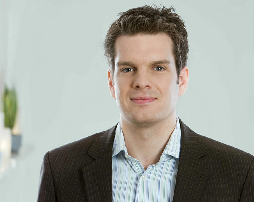 A man with short dark hair is wearing a pinstripe suit jacket and a light-striped shirt, standing in front of a plain light background, exuding the professionalism typical in the digital commerce industry.