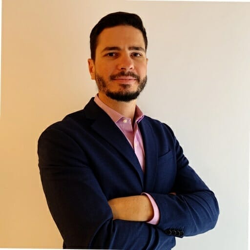 A man with short dark hair and a beard stands with arms crossed, wearing a navy blazer and pink shirt, looking confidently at the camera, embodying the professional energy of e-show Madrid.