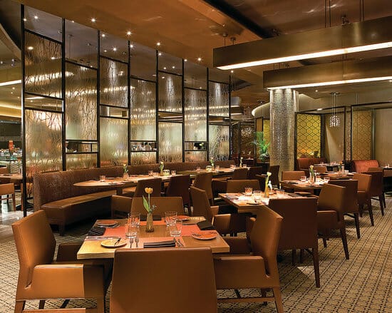 A modern restaurant interior with brown leather seating, wooden tables set with glasses and utensils, and decorative glass partitions. Seamless design and warm lighting create a cozy atmosphere that blends contemporary aesthetics with a hint of Saudi Arabian charm.