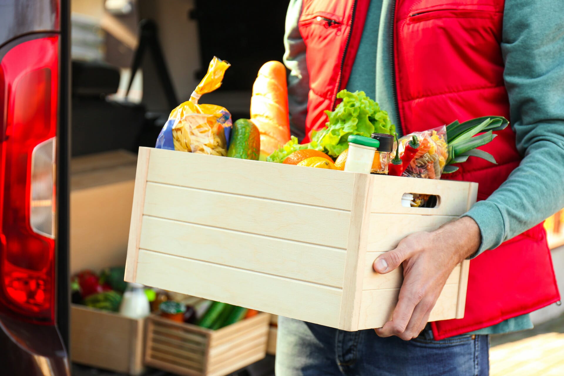 A person in a red vest is holding a wooden crate filled with assorted groceries from Aldi US, including bread, fresh vegetables, and packaged items.