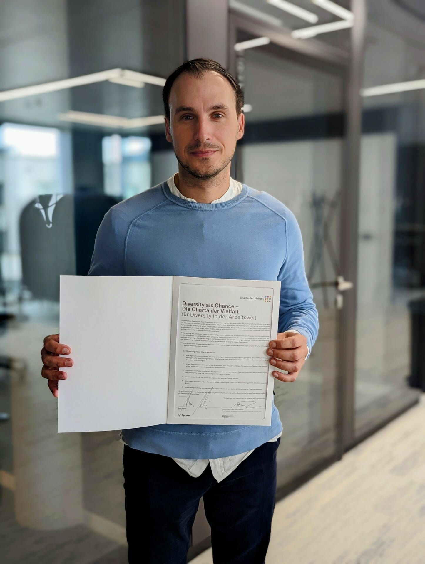 A person in a blue sweater holding an open document in a modern indoor setting with glass walls in the background, reflecting the commitment to sustainability.