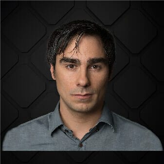 A man with dark hair and a neutral expression, wearing a gray collared shirt, stands in front of a geometric-patterned, dark background, embodying the modern professional in digital commerce.