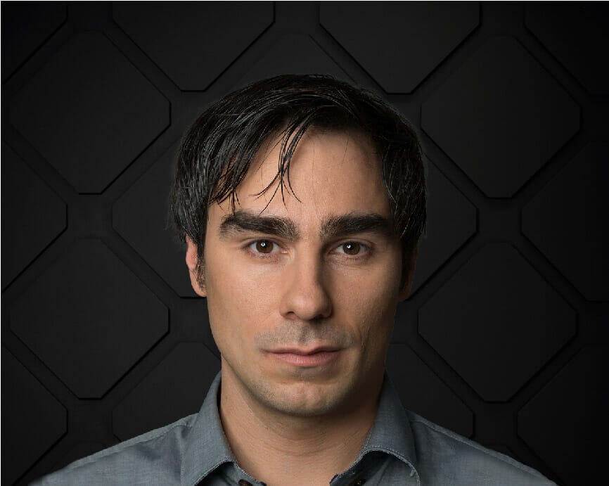 A man with dark hair and a serious expression is facing forward. He is wearing a gray shirt and standing against a dark, geometric-patterned background, subtly capturing the essence of digital commerce.