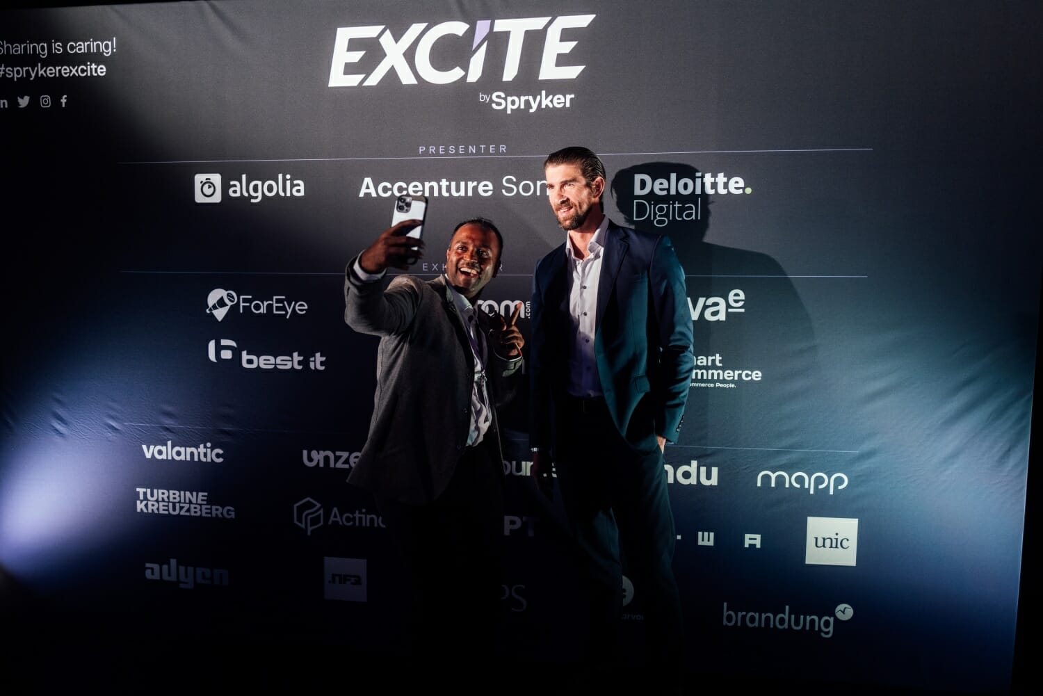 Two men in business attire take a selfie in front of a sponsorship backdrop at the "Spryker EXCITE 2023" event.