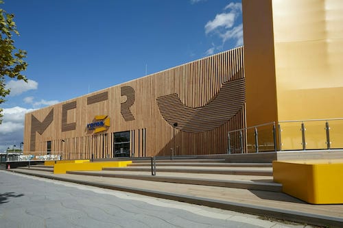 Modern building with wooden panel exterior and large decorative cutouts, featuring a sign that reads "NORR." The structure has glass railings and a staircase at the entrance, creating an inviting space for Spryker customers.