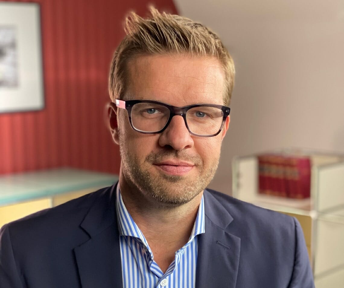 A man with blonde hair, glasses, and a suit is looking at the camera. He is in an indoor setting with a red wall and bookshelf in the background, possibly preparing for the 2023 Customer Advisory Board meeting.