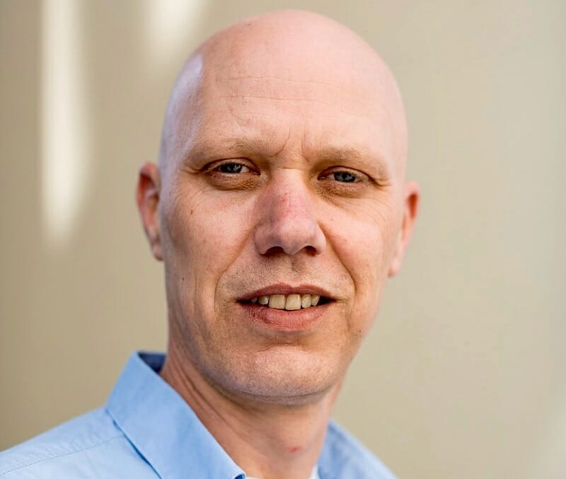 A bald man with a slight smile is wearing a light blue collared shirt, standing against a neutral background, possibly ready for the 2023 Customer Advisory Board meeting.