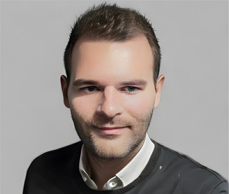 A digitally rendered portrait of a man with short hair, light facial hair, and a neutral expression, wearing a white collared shirt under a dark sweater, set against a plain background. Ideal for professional settings or 2023 Customer Advisory Board presentations.