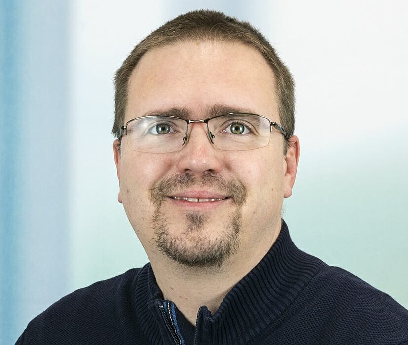 A man with short brown hair, glasses, goatee, and a navy blue zip-up sweater smiles at the camera against a blurred background during the 2023 Customer Advisory Board meeting.