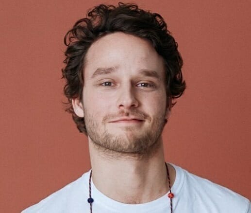 A person with curly hair and a short beard, wearing a white shirt and a beaded necklace, stands against a solid brown background, appearing as though they have just stepped out of an important customer advisory board meeting.