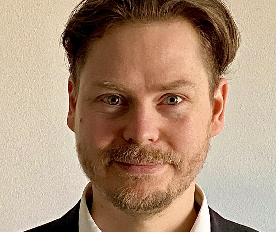 A man with short, brown hair, a beard, and mustache is wearing a white shirt and black jacket, looking straight ahead with a neutral expression at the 2023 Customer Advisory Board meeting.