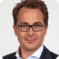 A man with brown hair and glasses wearing a dark suit jacket and white shirt poses for a professional headshot against a plain background, exemplifying the confidence and professionalism that Spryker customers value.