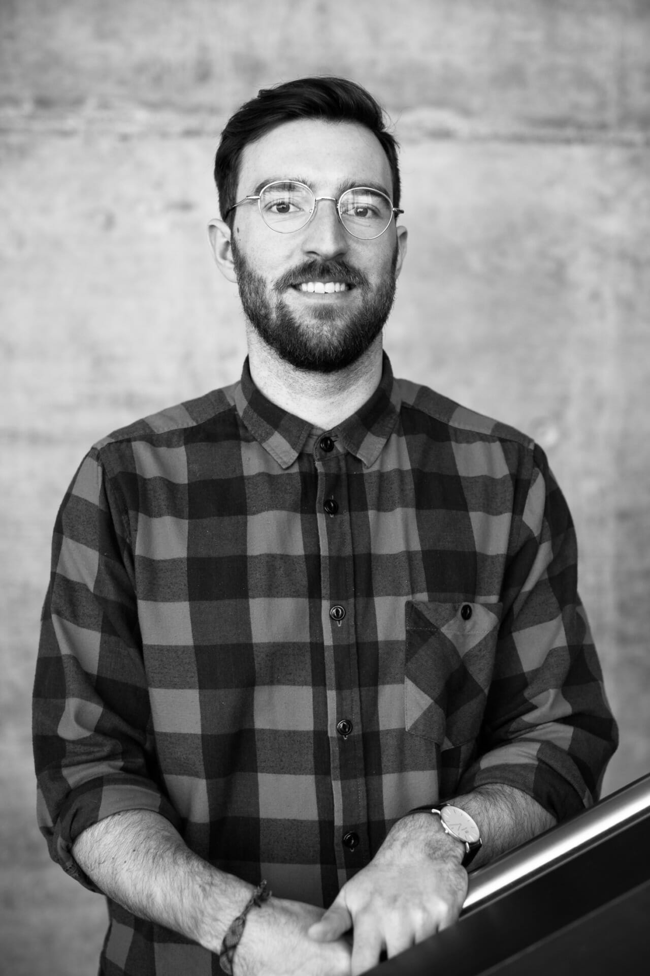 Black and white photo of a man standing with a slight smile, wearing glasses and a plaid shirt, his hands resting on a railing, as if presenting to the customer advisory board.