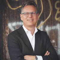 A man with glasses and a suit stands with his arms crossed in front of a graffiti-covered wooden background, embodying the confidence and dynamism of Spryker customers.