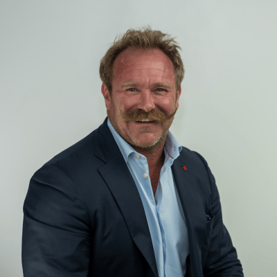 A man with a mustache is wearing a dark suit and light blue shirt, smiling at the camera against a plain background, preparing for Shoptalk Las Vegas 2023.