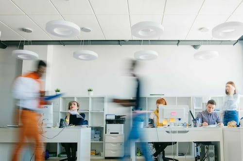 People working at desks in a modern, open-plan office with bright lighting. Some individuals are seated and using computers, while others are walking around. Spryker customers can be seen engaging in lively discussions. The background includes shelves and office supplies.