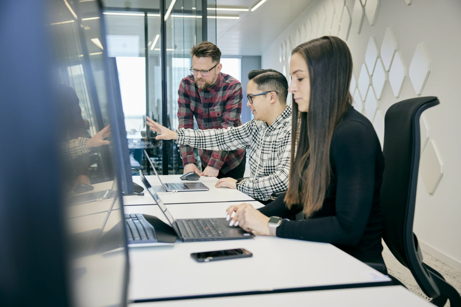 Three people work at a table with two laptops and a phone, immersed in delivering Composable Value Services. One person points at a screen while the others concentrate on their devices.