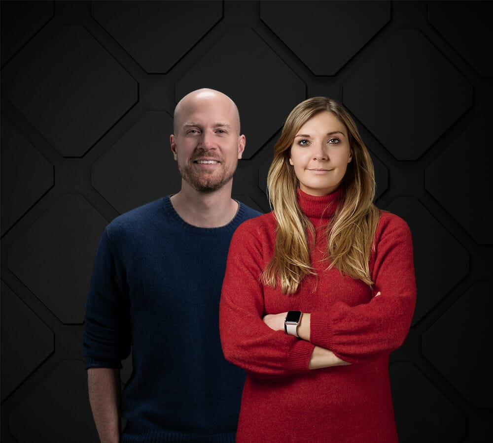 Two individuals, one wearing a navy blue sweater and another in a red sweater with long hair and a smartwatch, stand against a dark geometric background. The scene subtly evokes the efficiency of Spryker services in orchestrating seamless solutions.