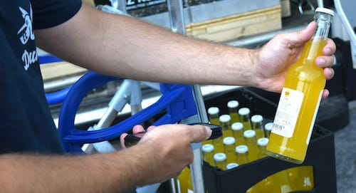 A Spryker customer scans the barcode of a glass bottle with a handheld scanner, while a crate of similar bottles waits in the background.