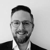 Black-and-white photo of a man with glasses and a beard, wearing a suit, smiling at the camera—capturing the essence of professionalism that appeals to Spryker customers.