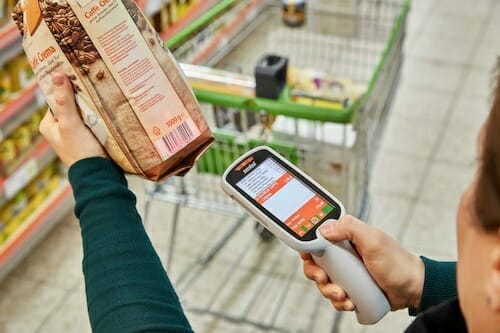 A spryker customer is seen scanning a bag of coffee beans with a handheld scanner in a grocery store, while a shopping cart waits in the background.