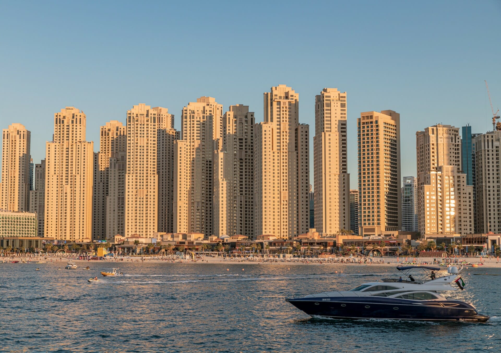 Tall beige skyscrapers tower over a bustling beach, with a sleek black yacht cruising on the water in the foreground under a clear blue sky, reminiscent of the innovative spirit you’d find at Seamless Middle East 2023.