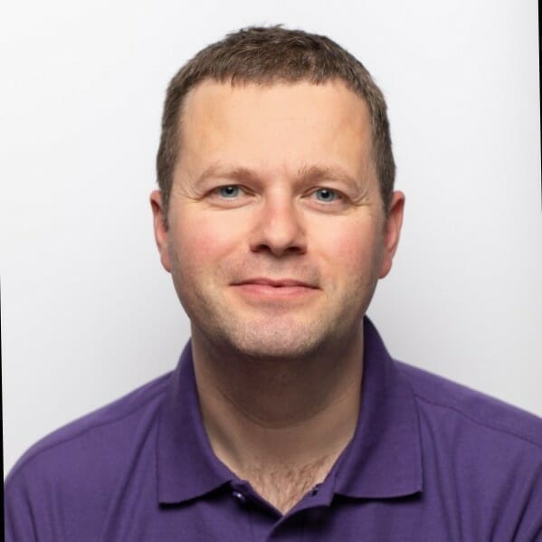 A man with short hair and blue eyes, wearing a purple polo shirt, stands against a white background, looking ready for the K5 Konferenz 2023.