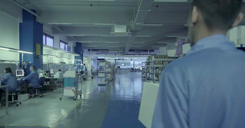 A person in a blue lab coat observes a spacious, well-lit laboratory with multiple workstations and other people working at computers in the background, meticulously analyzing industrial goods.