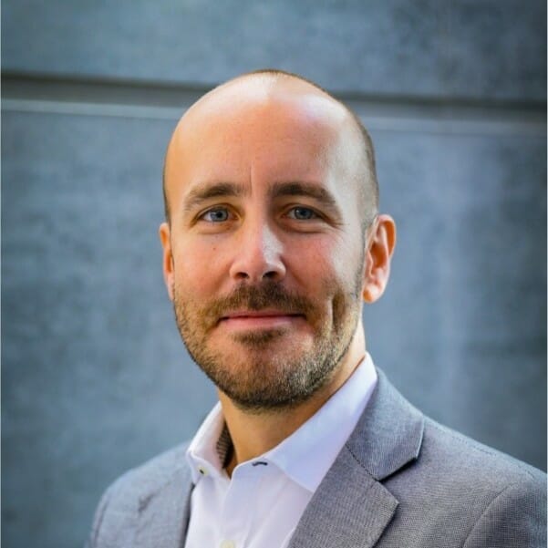 A bald man with a beard and mustache, wearing a light blue dress shirt and gray blazer, smiles slightly. The background is a plain gray wall, hinting at his participation in the K5 Konferenz 2023.