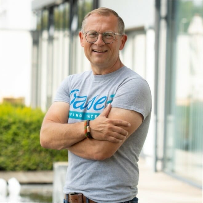 A person wearing glasses and a gray T-shirt with "Save" written on it stands outside with arms crossed, smiling at the camera, ready to attend the K5 Konferenz 2023.