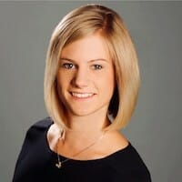 A woman with shoulder-length blonde hair and a black top smiles confidently at the camera, set against a grey background reminiscent of the sleek design often found in industrial goods.