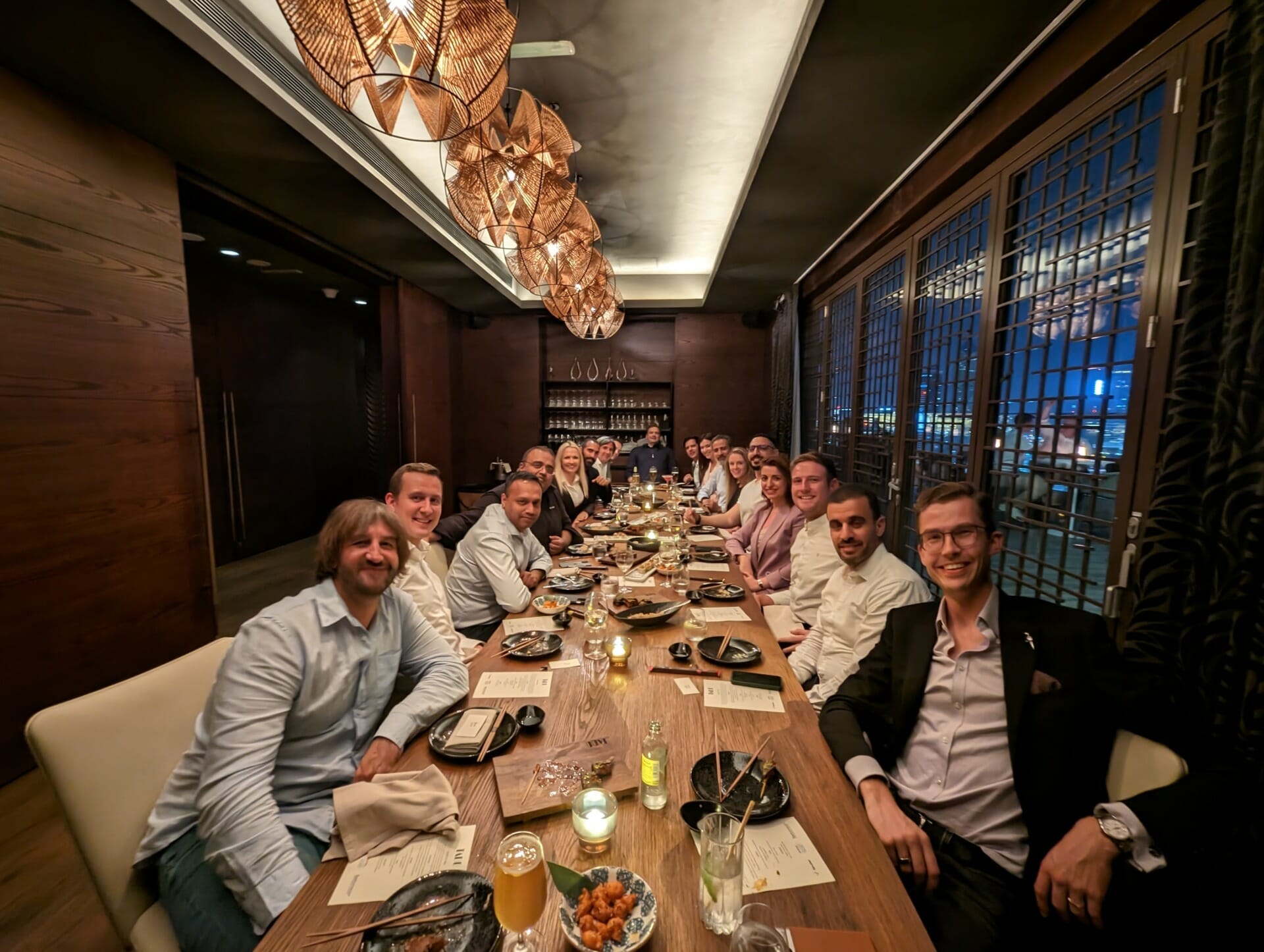 A group of people seated around a long table in a restaurant, engaging in conversation and dining. The ambiance is warm, with overhead pendant lights and wooden decor, reminiscent of the inviting settings found at Seamless Saudi Arabia 2023.