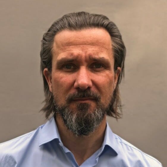 A man with shoulder-length hair and a beard, wearing a light blue collared shirt, stands against a plain background, looking like he could be discussing digital commerce strategies for manufacturers.