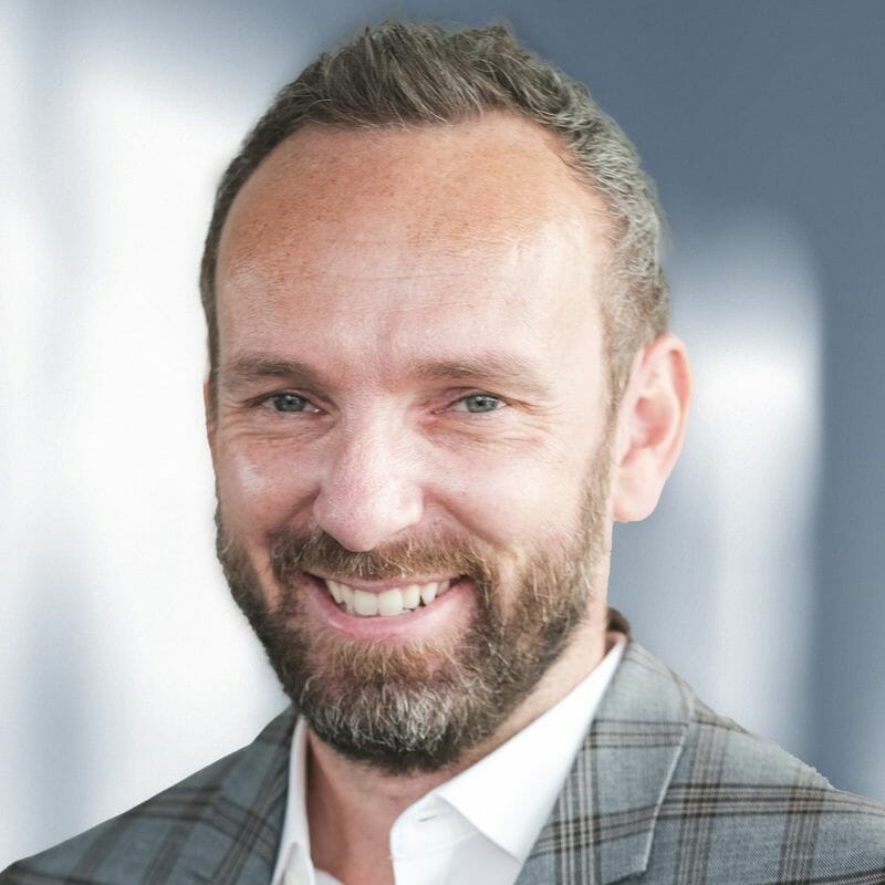 A man with short hair and a beard, smiling, is wearing a plaid jacket. The background is blurred, hinting at his focus on digital commerce strategies for manufacturers.