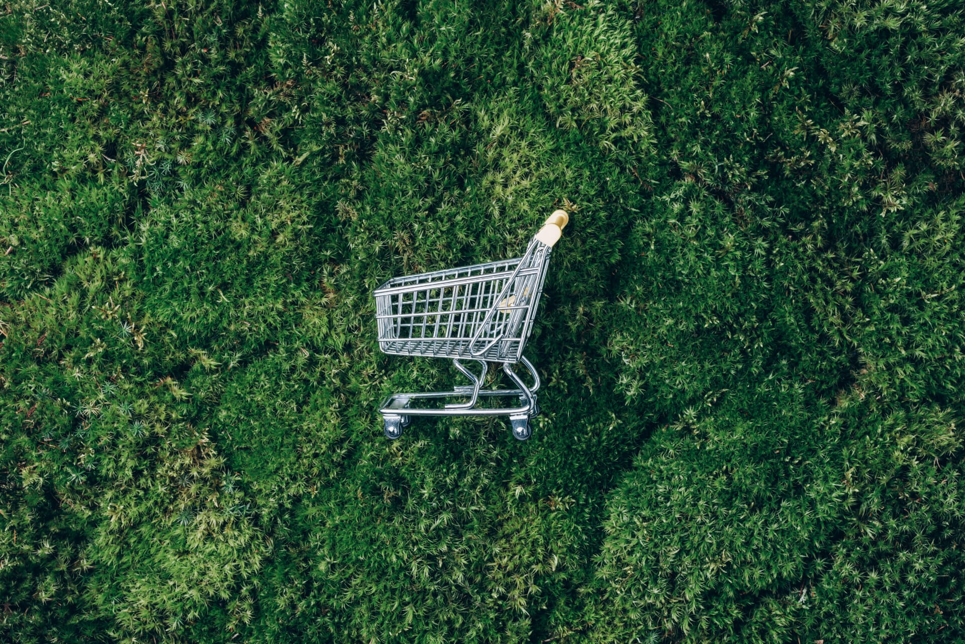 A small, empty shopping cart sits on a green, grass-like surface, symbolizing the shift toward sustainable commerce.