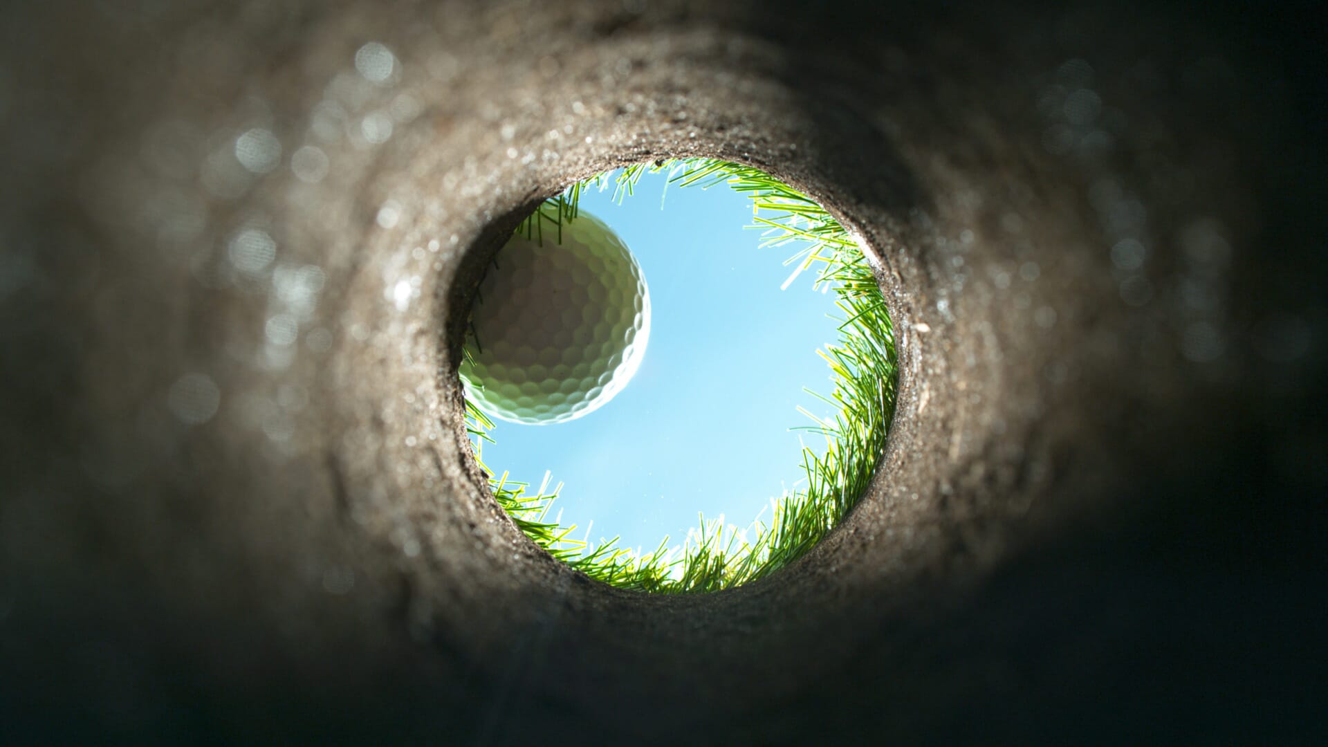 View from inside a golf hole looking up at a golf ball on the edge, with grass and blue sky visible in the background—a perfect moment to reflect on how sustainable commerce can help keep our greens pristine.