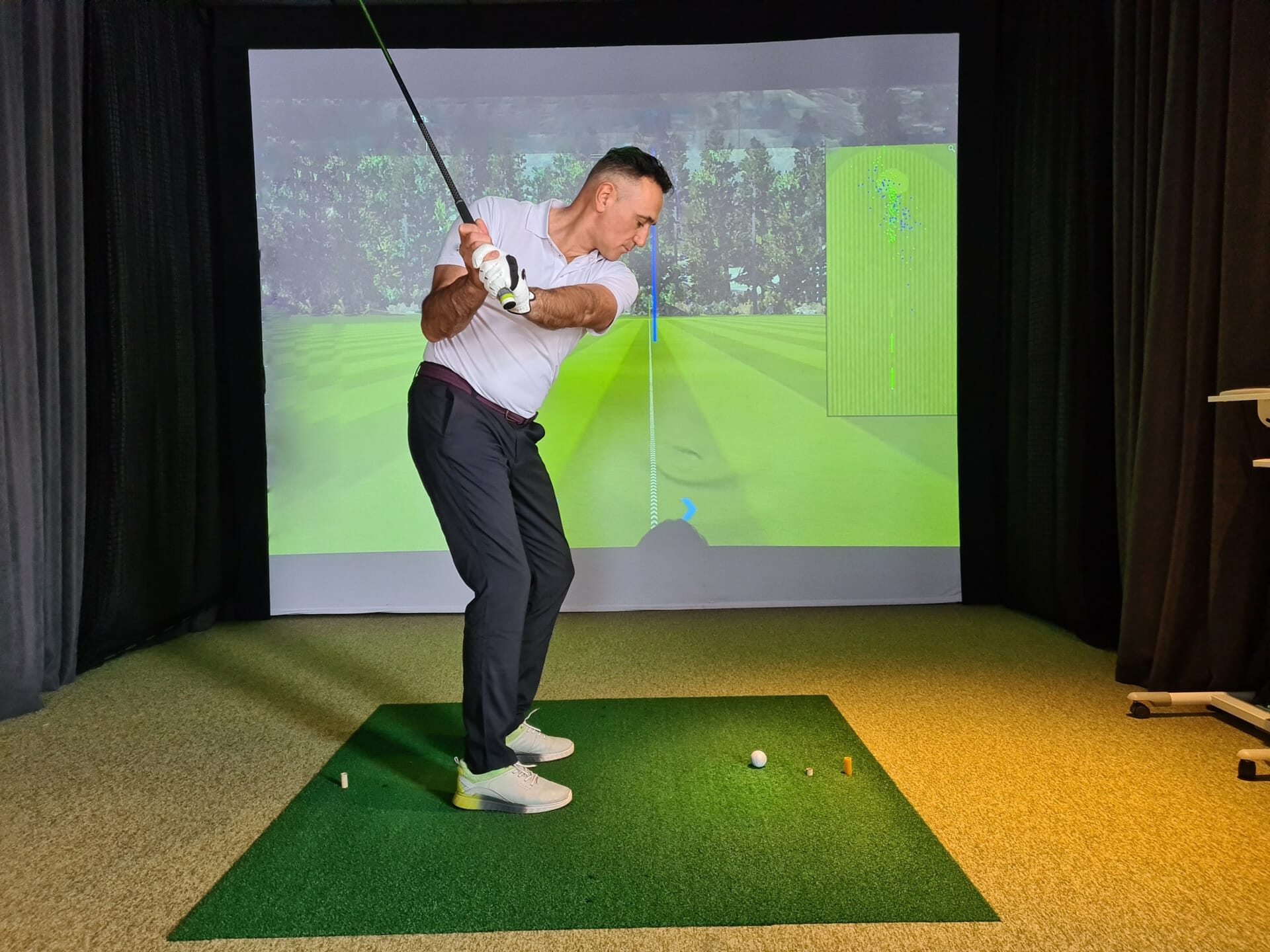 A man in a white shirt and dark pants is in mid-swing at an indoor golf simulator. He stands on a green mat with practice tees and a ball, with a screen displaying a golf course behind him, epitomizing the modern blend of sports and sustainable commerce.