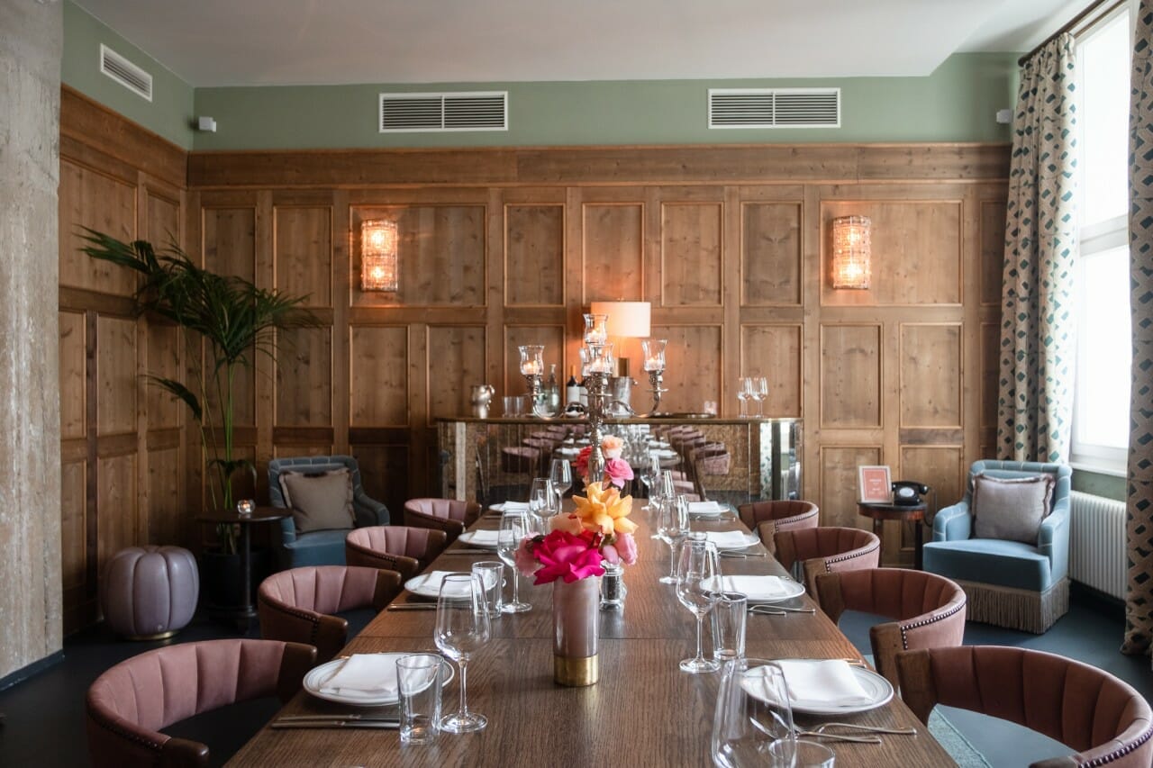 A dining room with a long wooden table set for a meal, surrounded by chairs. The room has wood paneling, potted plants, and decorative lights. A vase of flowers and glassware are on the table, echoing a hint of the future of grocery with fresh produce seamlessly integrated into elegant decor.