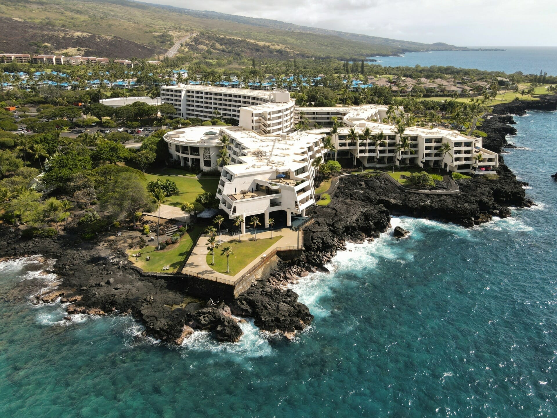 Aerial view of a large white coastal resort perched on black volcanic rocky cliffs, surrounded by lush greenery and with a blue ocean in the foreground. Buildings and a hillside can be seen in the background, reminiscent of the retreats frequented by B2B Florida manufacturers and distributors.