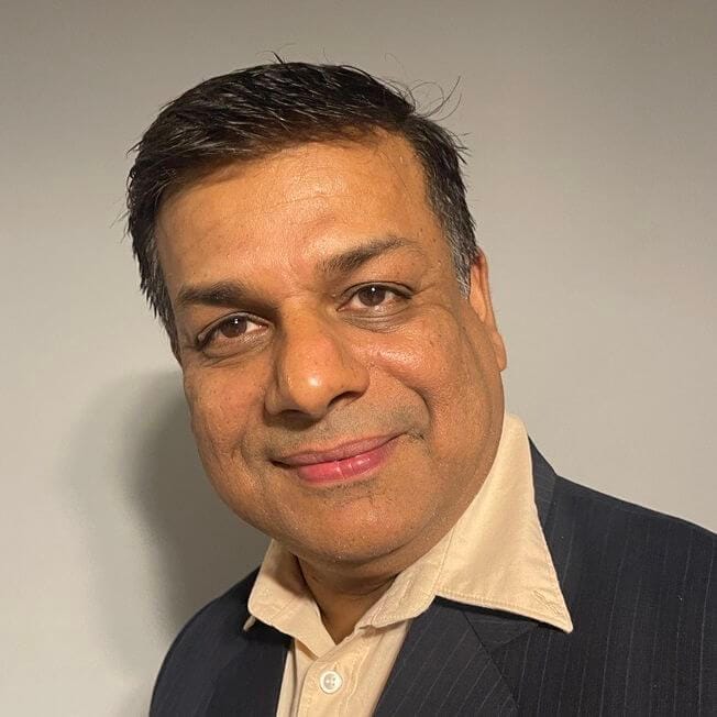 A man with short dark hair, wearing a light-colored shirt and dark suit jacket, smiling at the camera against a plain background, epitomizing the forward-thinking approach embedded in digital transformation strategies for manufacturers.