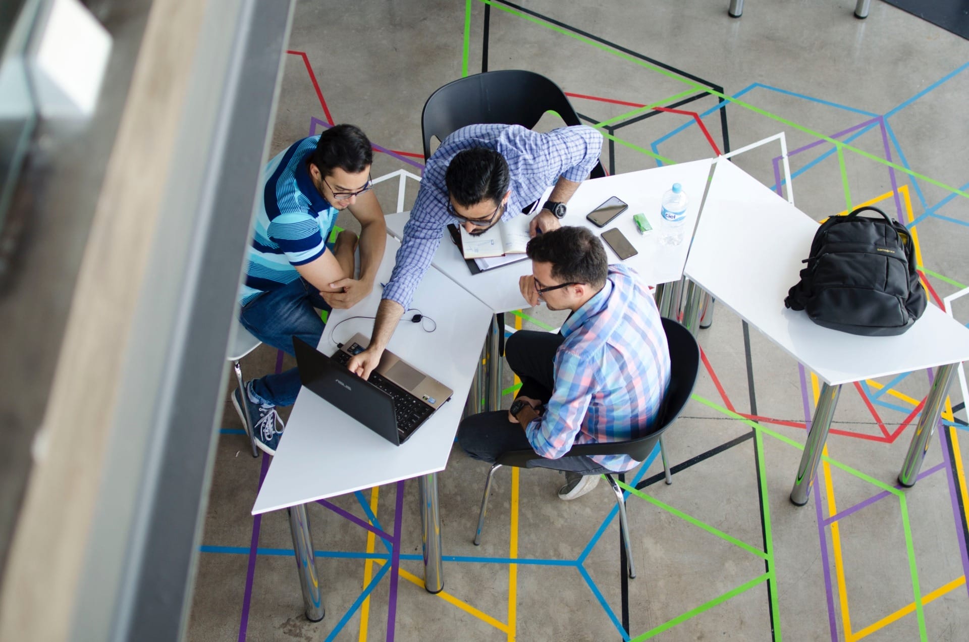 Three people sit around a white table, working on laptops and taking notes. The ground features colorful geometric lines, and a backpack is placed on an adjacent table. This workspace is perfect for B2B online meetings between European manufacturers and distributors.