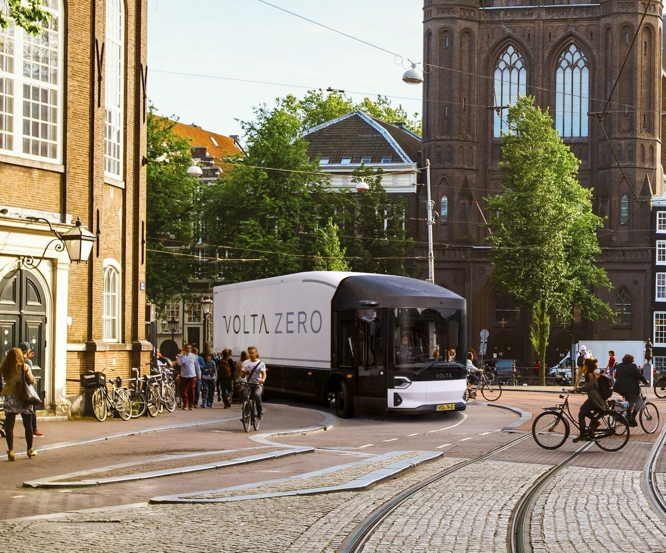 A Volta Zero electric truck drives through a European city street near a large historic building as pedestrians walk and cyclists ride nearby, seamlessly integrating technologies from Spryker and AWS to ensure efficient, sustainable performance.