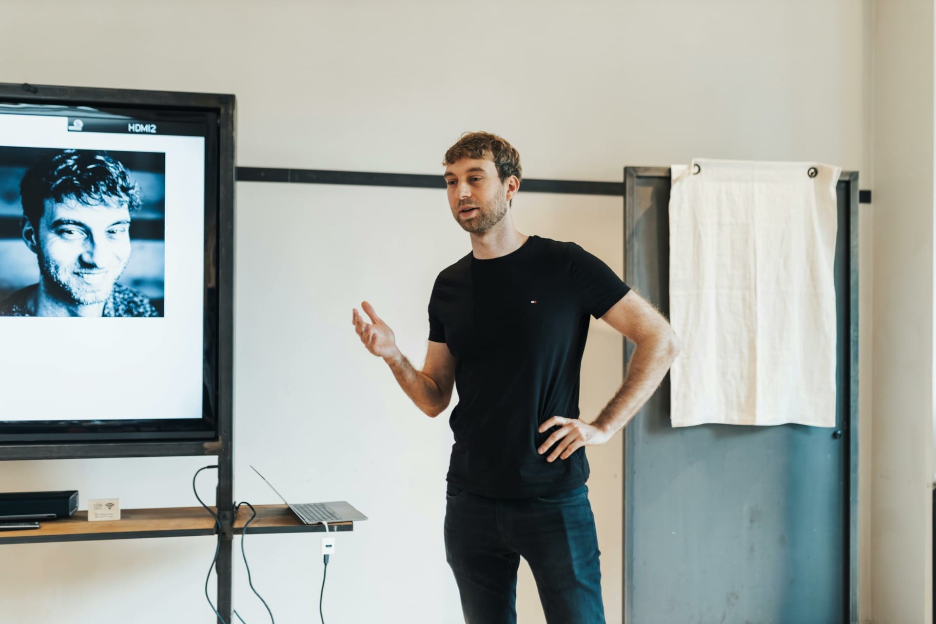 A person wearing a black shirt stands in front of a screen displaying a black and white photograph, giving a presentation on the innovations showcased at CES 2024. A laptop and a small object are on a table nearby.