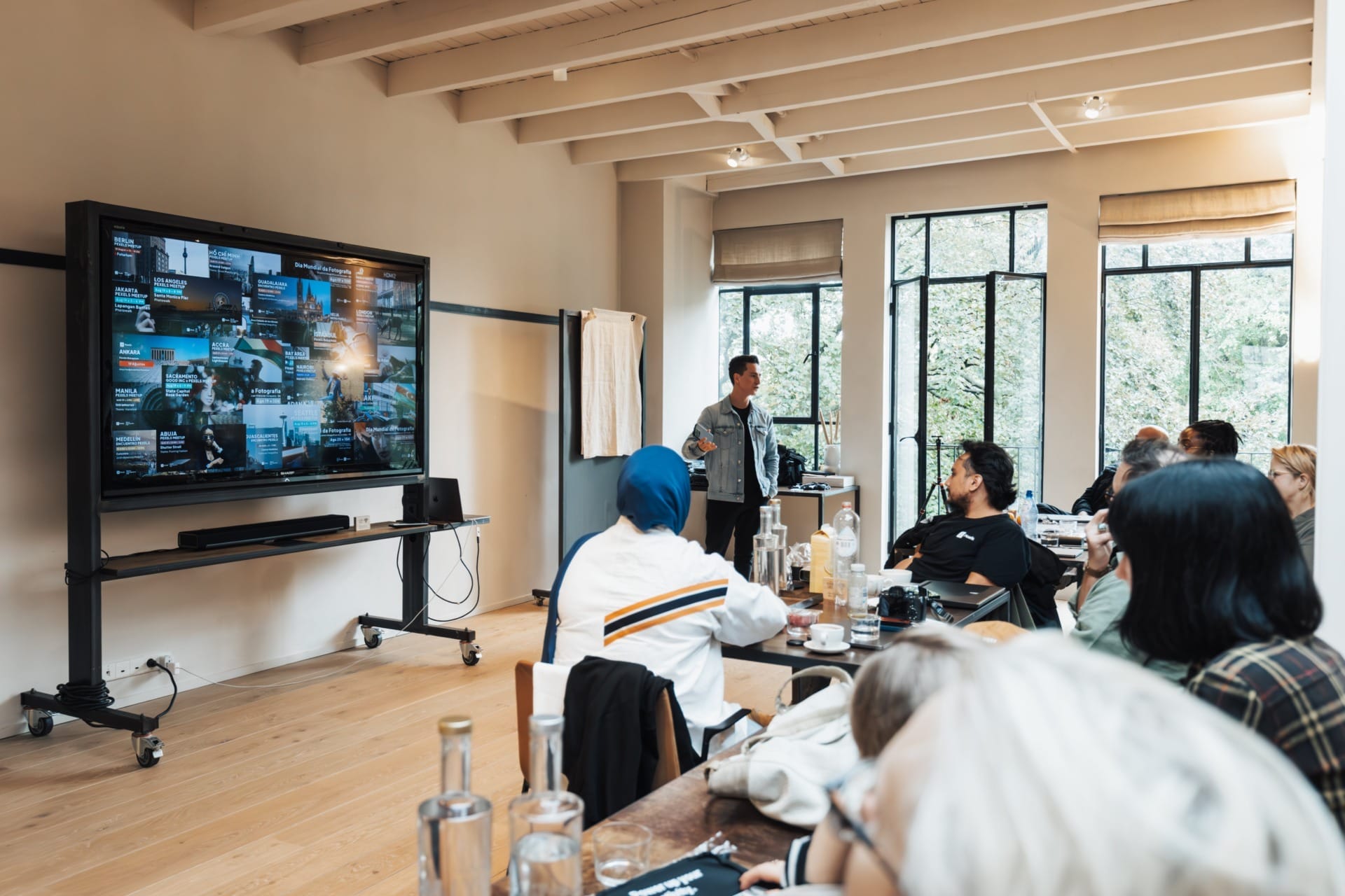 A person is giving a presentation to a seated audience using a large monitor displaying various images and information, reminiscent of CES 2024. The room has large windows and several tables with items on them.