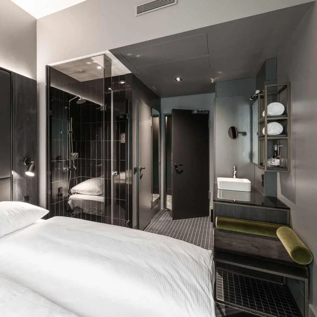 A modern, minimalist hotel room with a bed in the foreground, black walls, and contemporary decor. The bathroom features black tiled walls, a glass shower partition, and a sink with a mounted mirror and shelves stocked with towels.