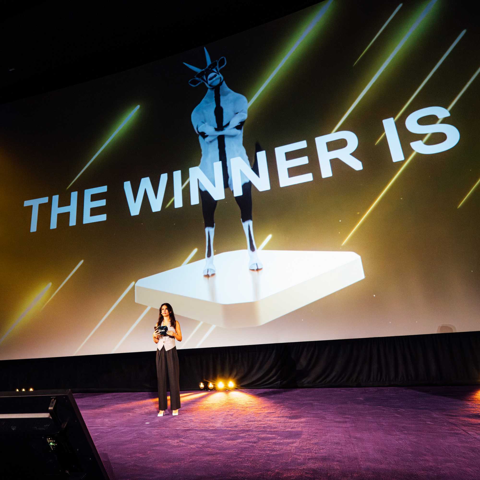 A woman stands on a stage with a microphone in hand, addressing an audience. Behind her on a large screen is an image of a goat standing on a pedestal with the text "THE WINNER IS" in bold letters. The stage is dimly lit with yellow spotlights.