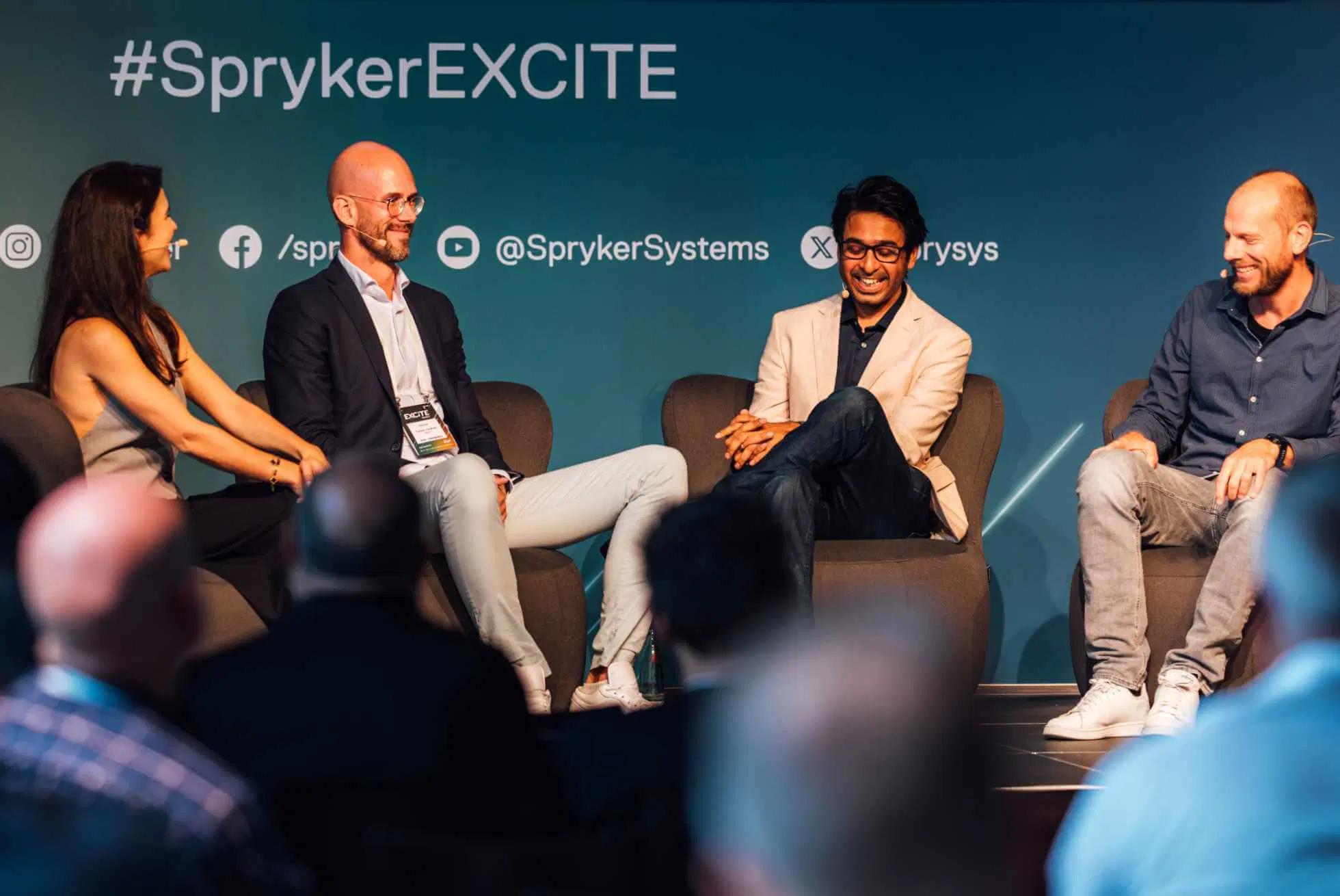 Four speakers are seated and engaged in discussion on stage at an event. The background has the hashtag #SprykerEXCITE and social media handles for SprykerSystems. Audience members are visible in the foreground. Everyone appears to be smiling and enjoying the conversation.