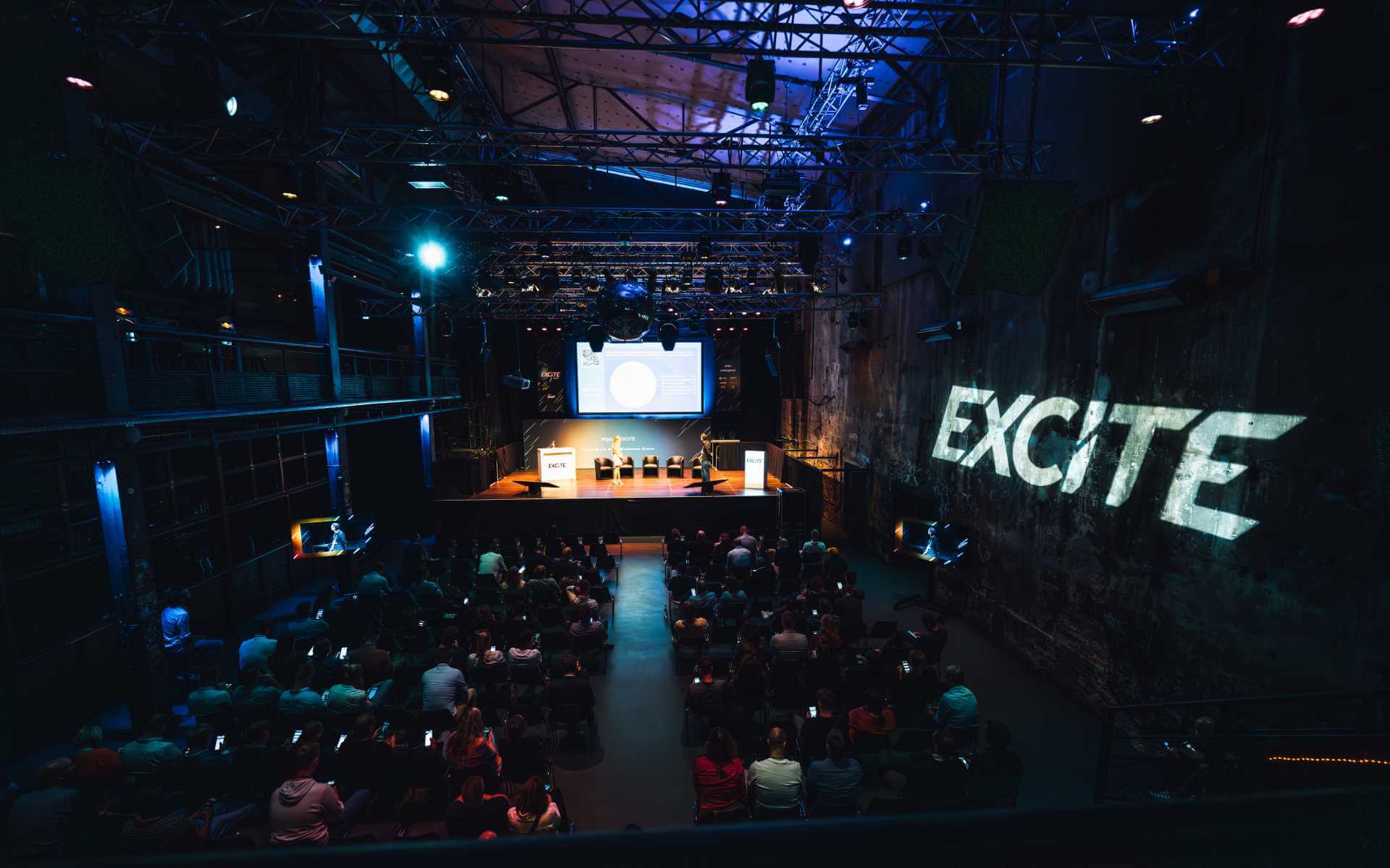 An audience is seated in a dimly lit auditorium, facing a stage with a large screen displaying a presentation. The word "EXCITE" is projected in bright light on the side wall. The stage showcases a few chairs and a podium. The ceiling features an array of lighting fixtures.