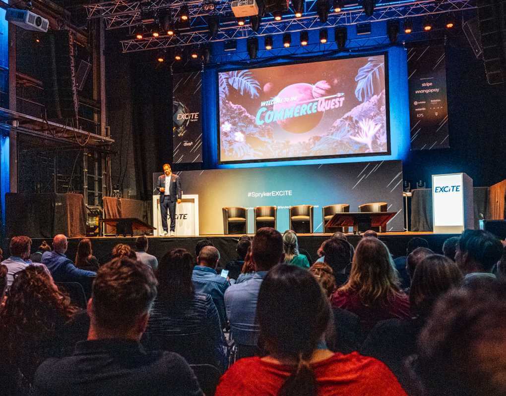 A person stands on a stage giving a presentation to an audience. Behind them is a large screen displaying "Commerce Quest" with a space-themed background. Several chairs are arranged on the stage, with branding and spotlights overhead.